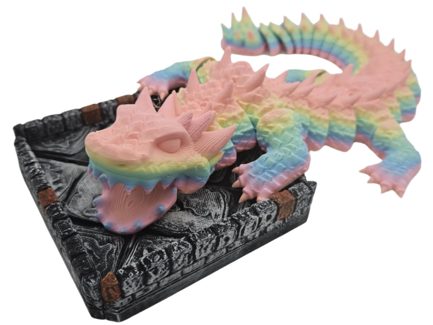 A rainbow pastel dragon is perched with its forelimbs on the edge of a gray, stone-patterned box, its head and tail extending away from the container.