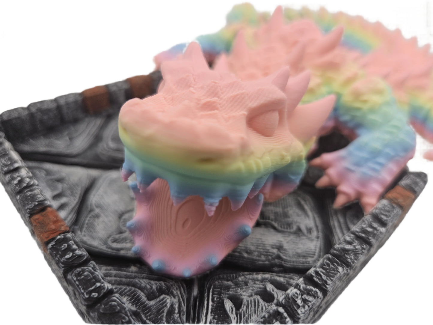 A rainbow pastel-colored, spiky dragon rests on a gray platform that looks like stone tiles with a brick border. The dragon’s mouth is wide open.