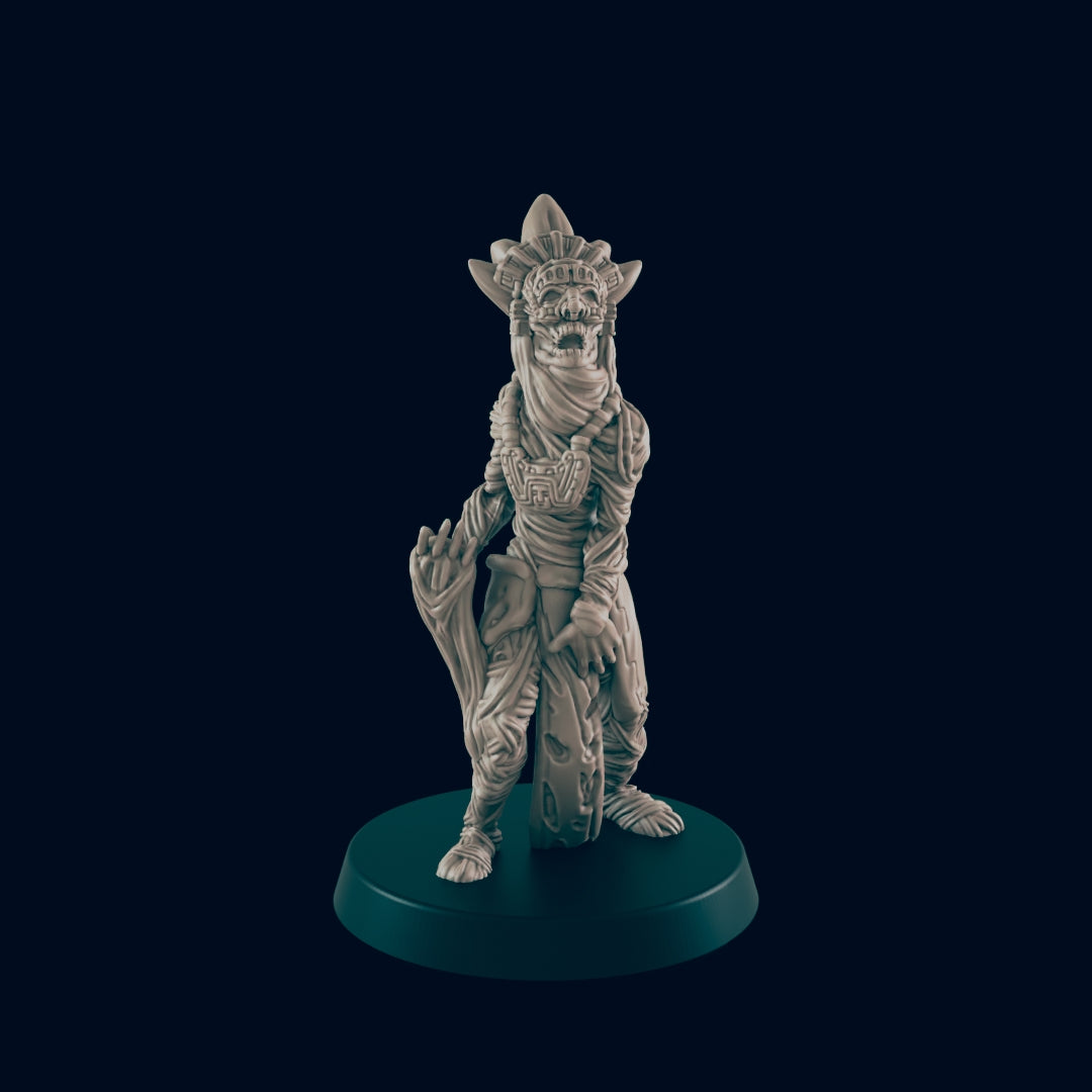 A skeletal, humanoid figurine stands on a circular base; it wears cloth wrappings and headdress, holding a circular object. The figurine is displayed against a dark background.