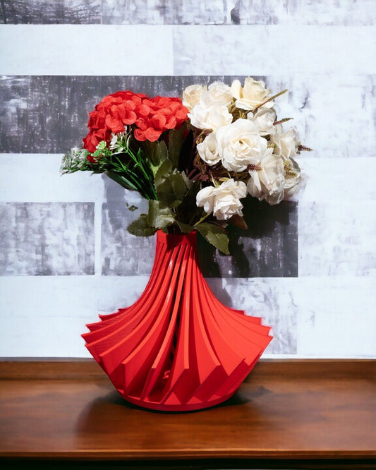 Flowers are arranged in a red, modern vase on a wooden surface, with a gray and white horizontally-striped patterned wall as a backdrop.