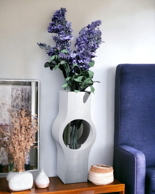 Purple flowers sit in a white vase with a circular opening. The vase is on a wooden table alongside various decorative items, set against a neutral wall and a blue chair.