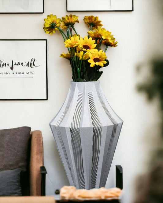A gray, textured vase holds yellow sunflowers. It sits on a surface near a brown chair, against a white wall adorned with framed artwork.  The artwork includes calligraphy that appears to read “_tam giác_” and more illegible text below.