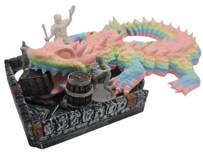 A rainbow dragon miniature is perched atop a dungeon wall set piece with miniatures, a barrel, and a treasure chest within.