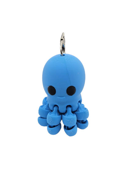 A small, blue, 3D-printed octopus keychain with black eyes and a subtle smile dangles from a silver keyring, against a black background.