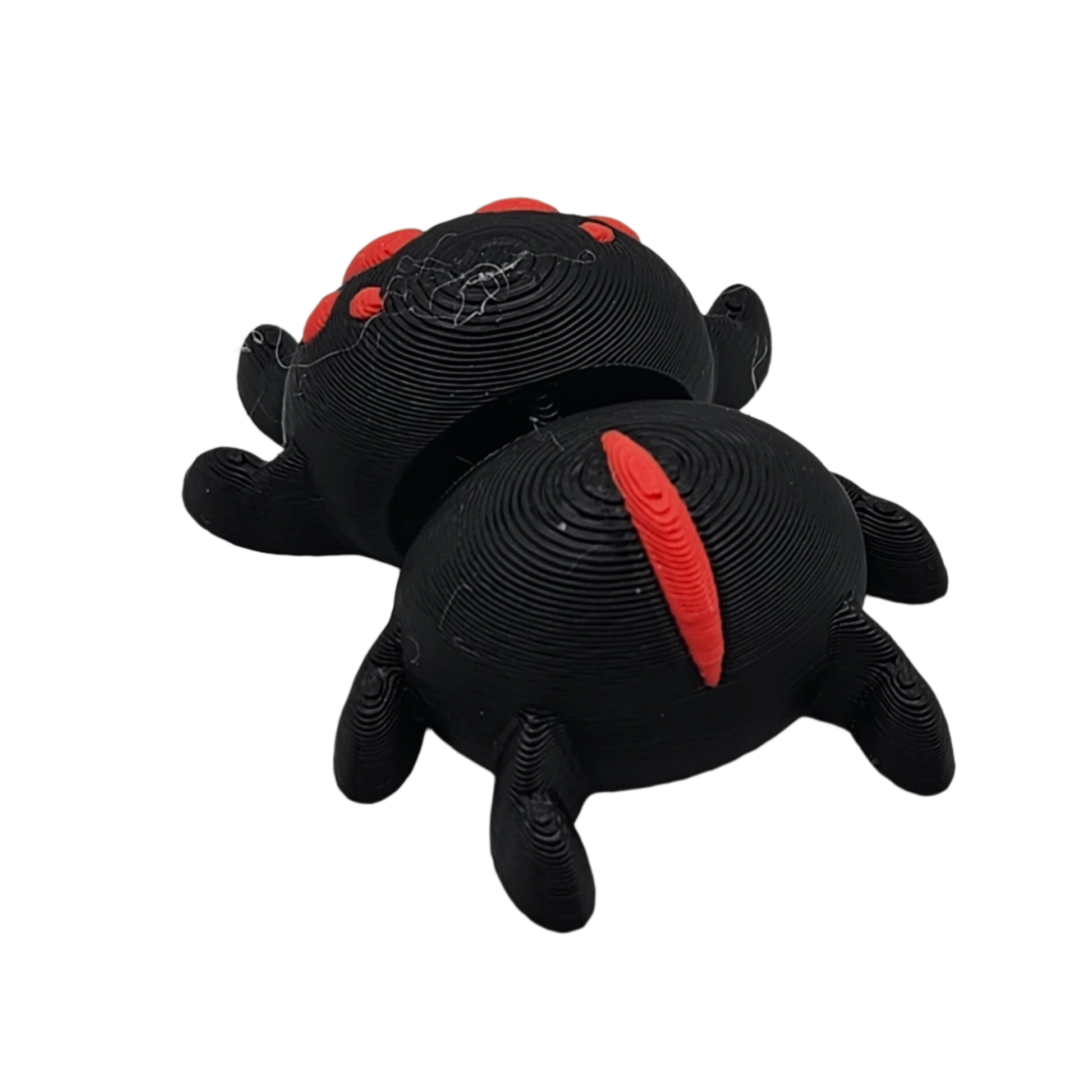 A black, 3D-printed toy spider lies on its back with red details. Its segmented body and legs are clearly visible against the plain background.