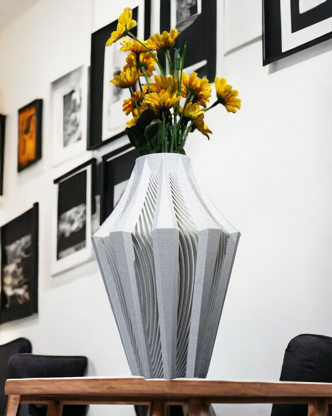 A light gray, intricately carved vase holds yellow flowers. It sits on a wooden table against a wall adorned with framed black and white photographs.