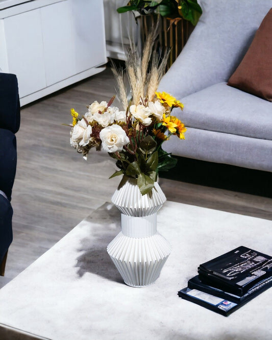 A white vase holds yellow, white, and brown flowers. The vase is on a coffee table next to some books, with a sofa in the background.