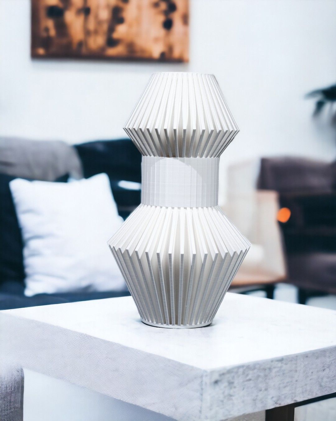 A white, vertically ridged, hourglass-shaped vase sits on a white table. Furniture and artwork are visible in the background, out of focus.