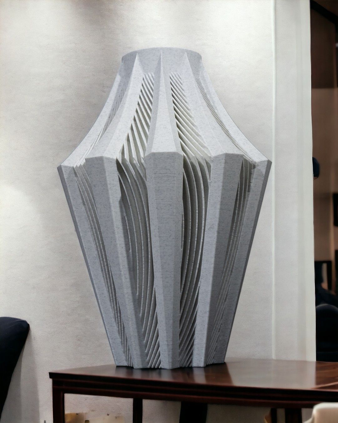 A large, light grey, intricately carved vase sits on a dark brown wooden table against a light wall.