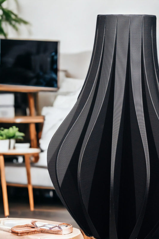 A tall, dark vase with fluted sides sits on a light-colored surface. Behind it, there is a blurred image of a television, shelves, plants and furniture.