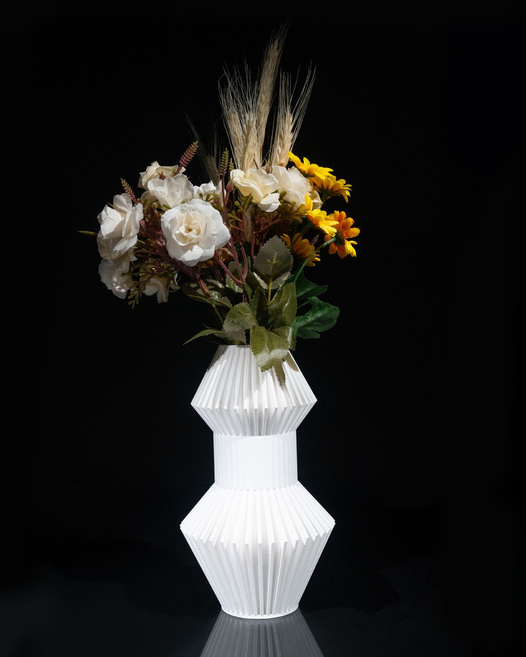 Flowers sit in a white, faceted vase. The arrangement includes white roses, yellow daisies, and wheat stalks, all against a dark, reflective background.