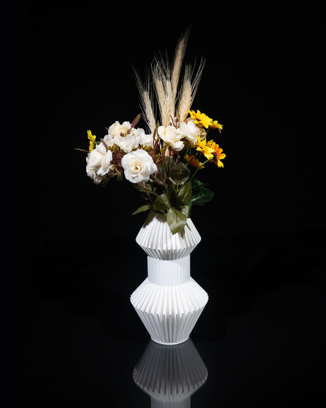 A white, hourglass-shaped vase holds a bouquet of white roses, yellow daisies, and wheat stalks. The vase sits on a shiny black surface, reflecting its image.