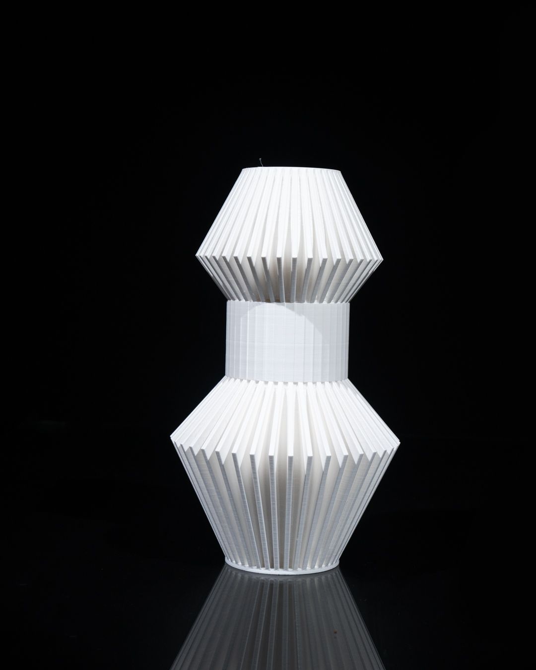 A white, vase-like object sits centered in a black space. The object is made of vertically ribbed pieces connected in a hourglass shape.