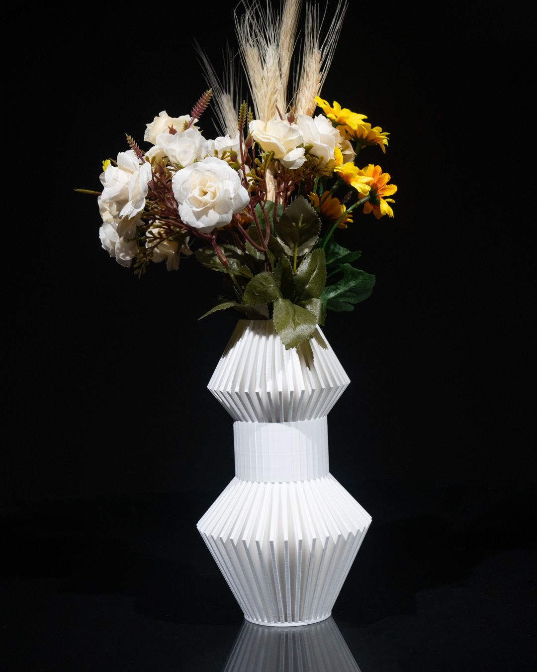 A white geometric vase holds a bouquet of white roses, yellow flowers, and wheat stalks against a dark background. The vase reflects on a black surface.