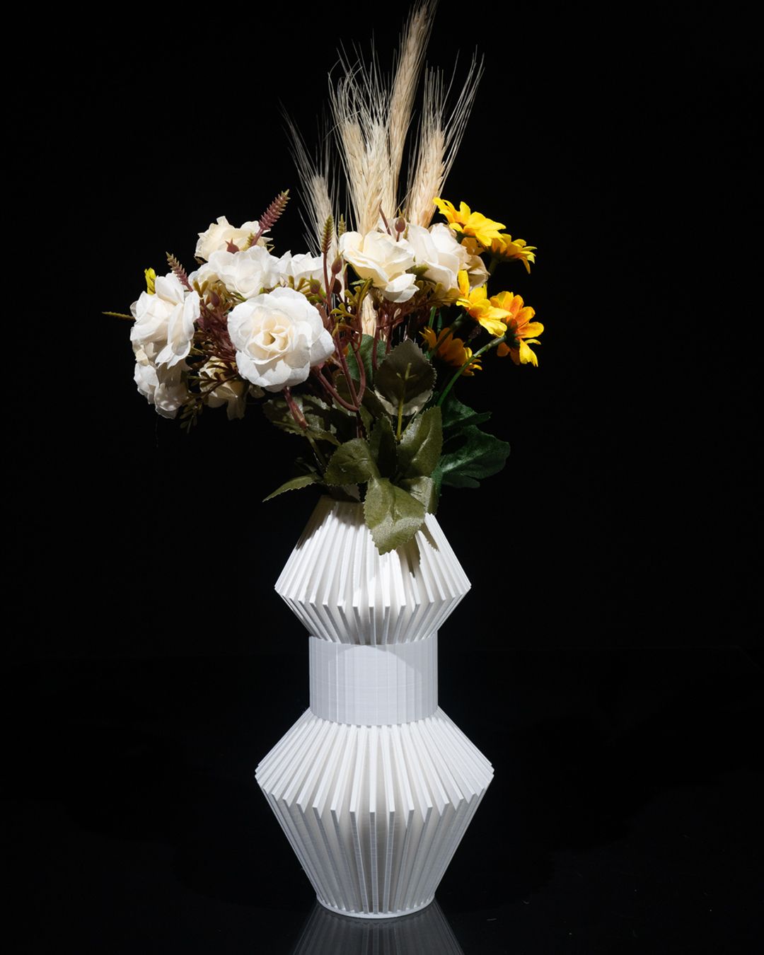 Flowers sit in a white, geometric vase, centered against a black backdrop. The bouquet includes roses, yellow flowers, and tall stalks of wheat.
