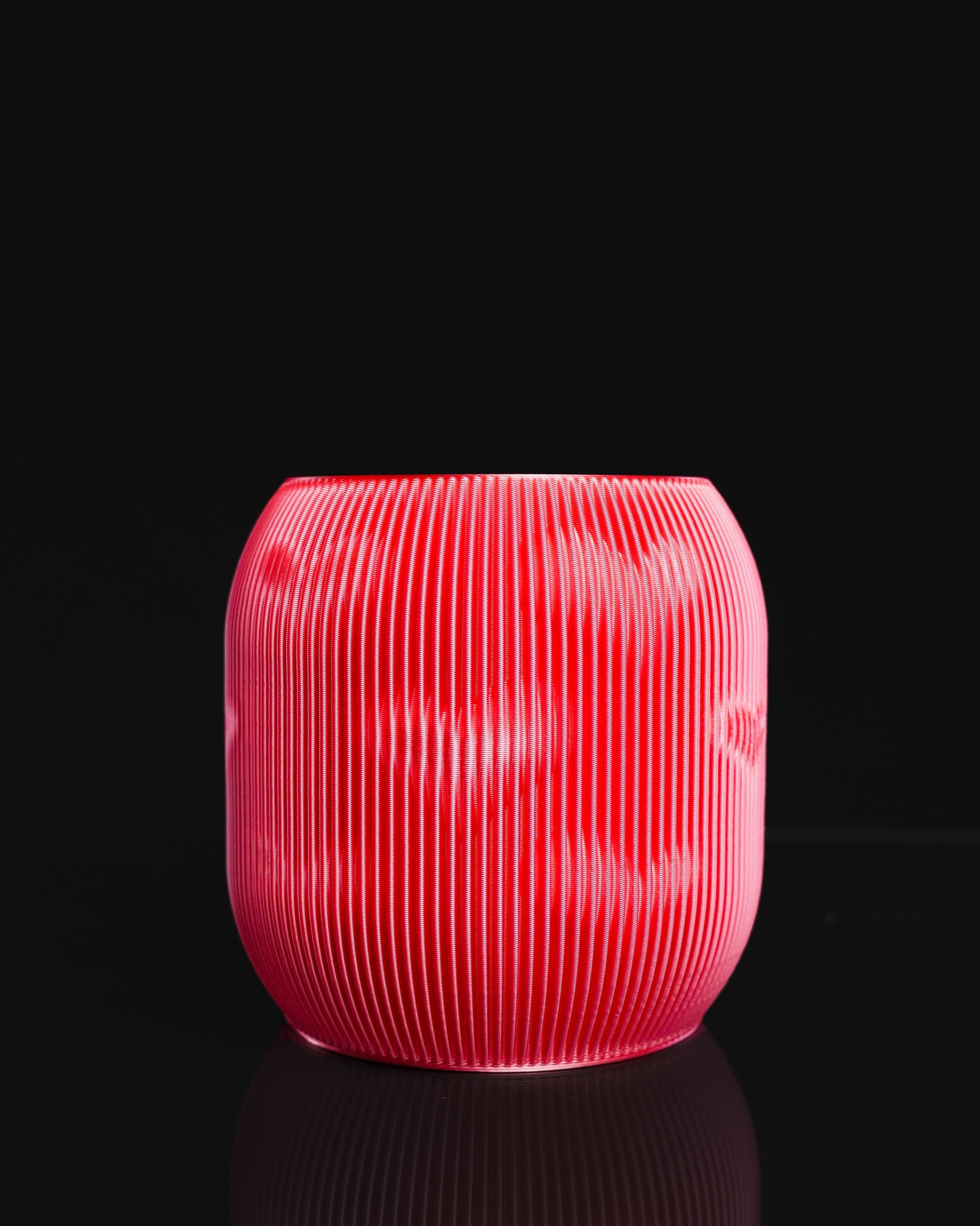 A red, vertically ribbed, vase-shaped object sits, reflected, on a black surface. It is set against a completely black background.
