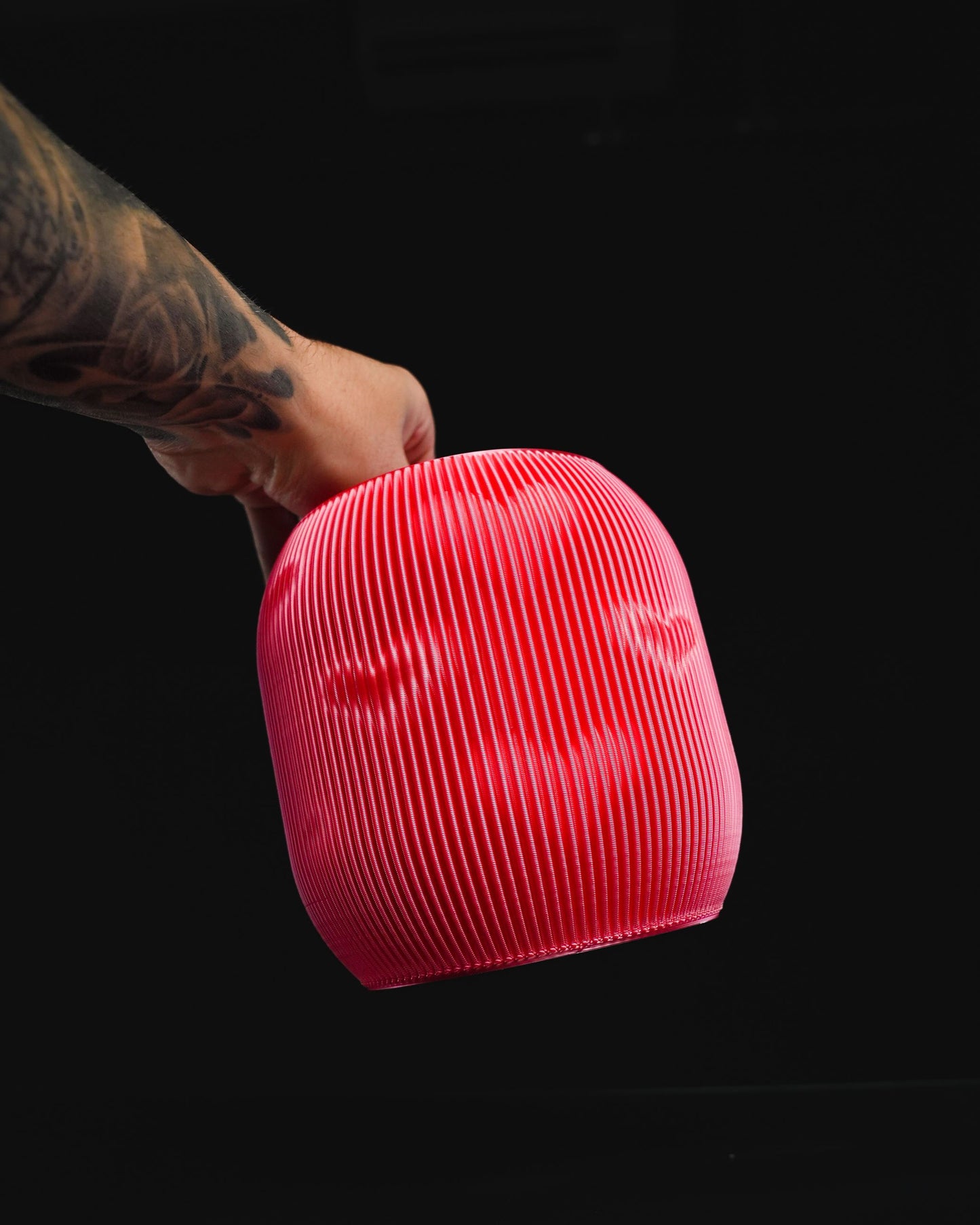 A tattooed hand holds a red, ridged, hollow, dome-shaped object. The object appears to be a lampshade. The background is solid black.