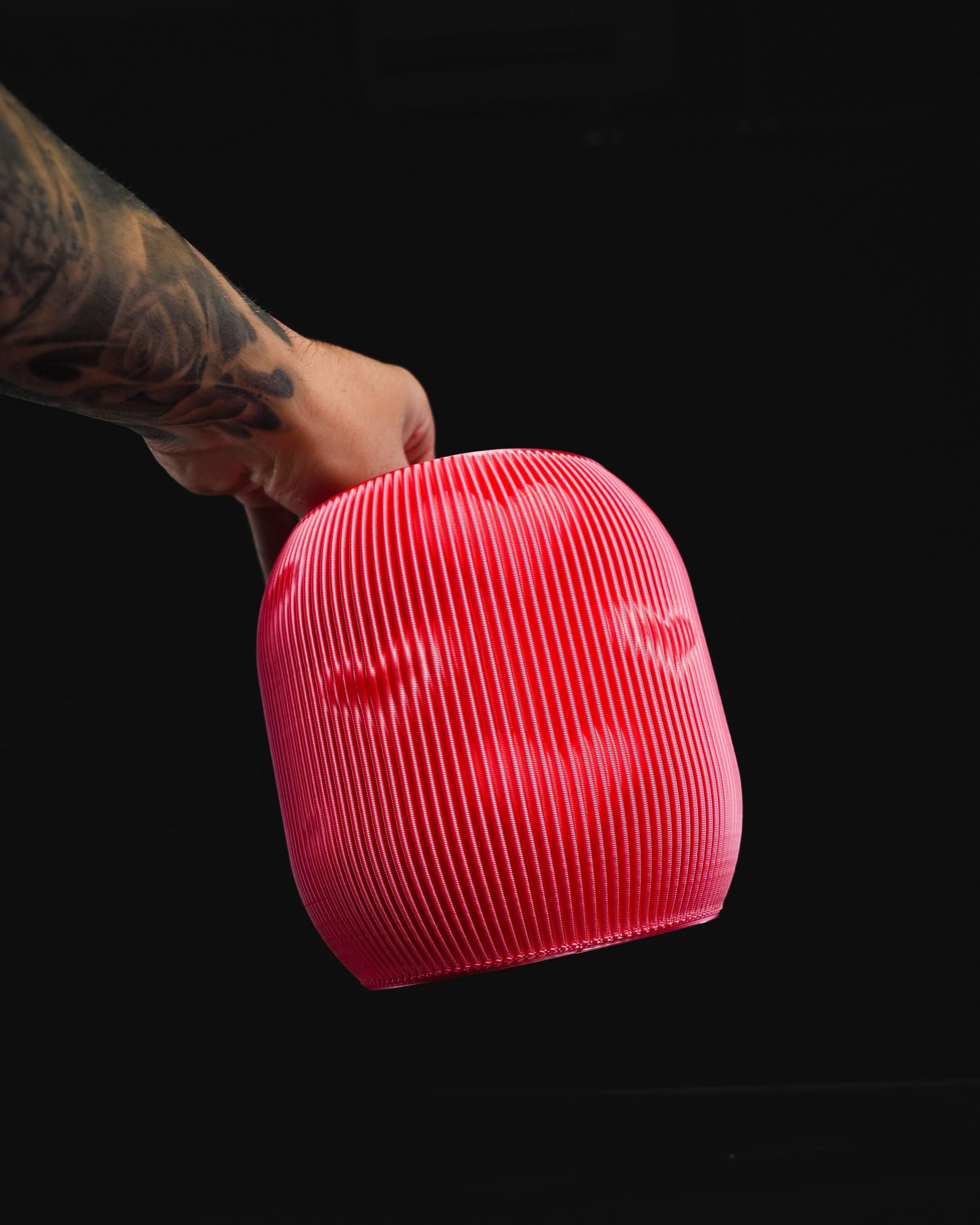 A tattooed hand holds a red, ridged, hollow, dome-shaped object. The object appears to be a lampshade. The background is solid black.