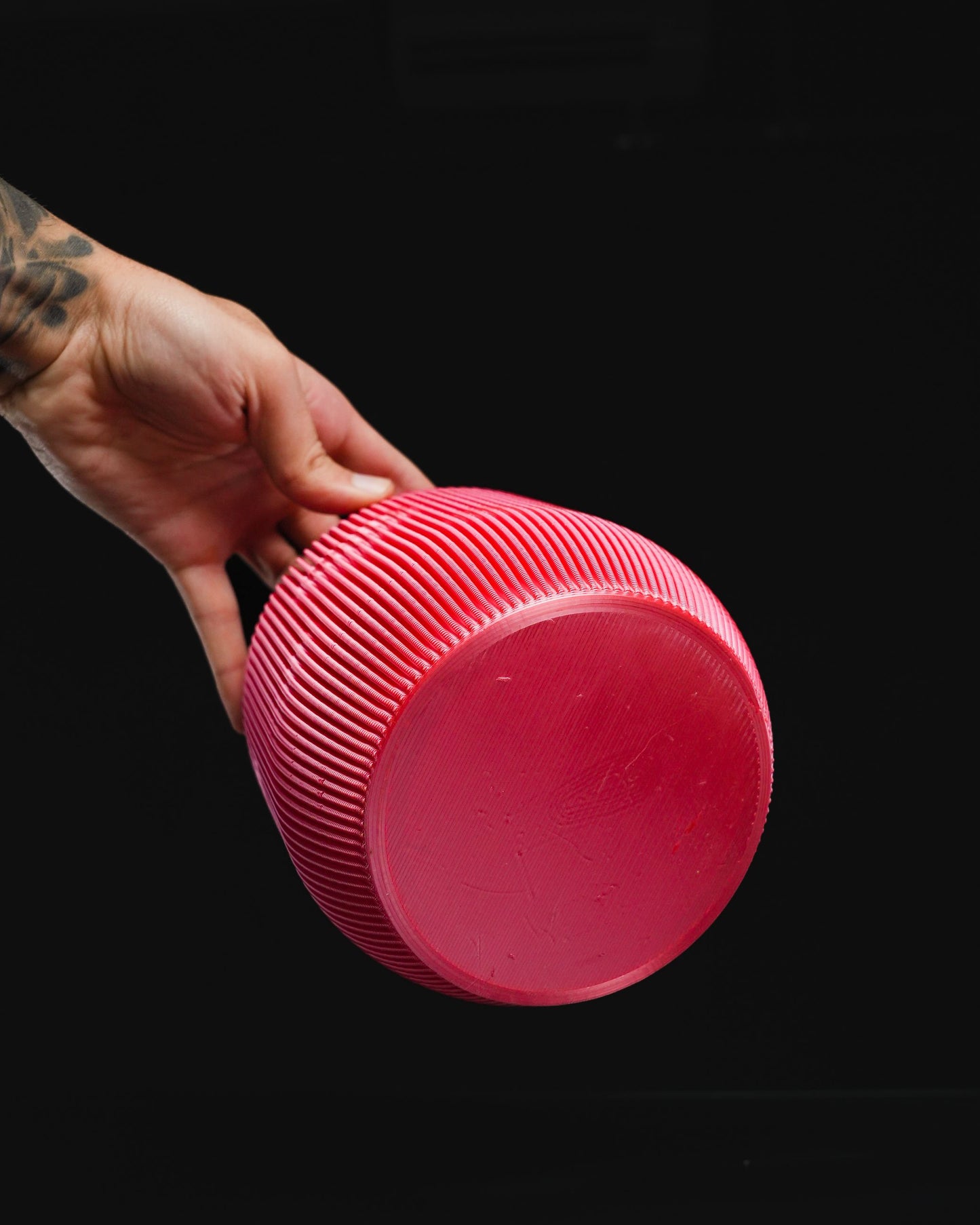A tattooed hand holds a pink, spherical object with parallel ridges against a solid black background.