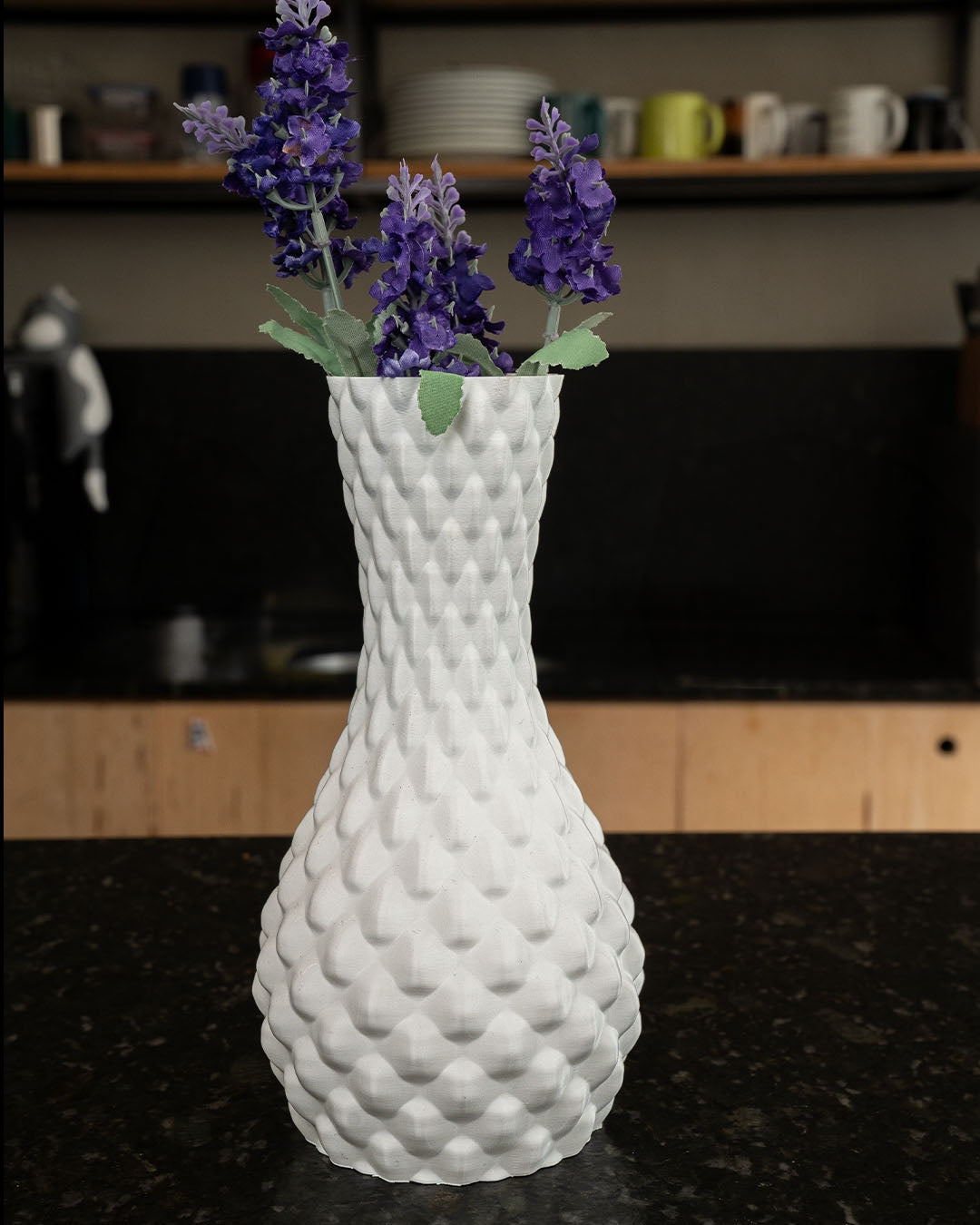 A textured white vase holds purple lavender; it sits on a dark countertop in a kitchen setting.