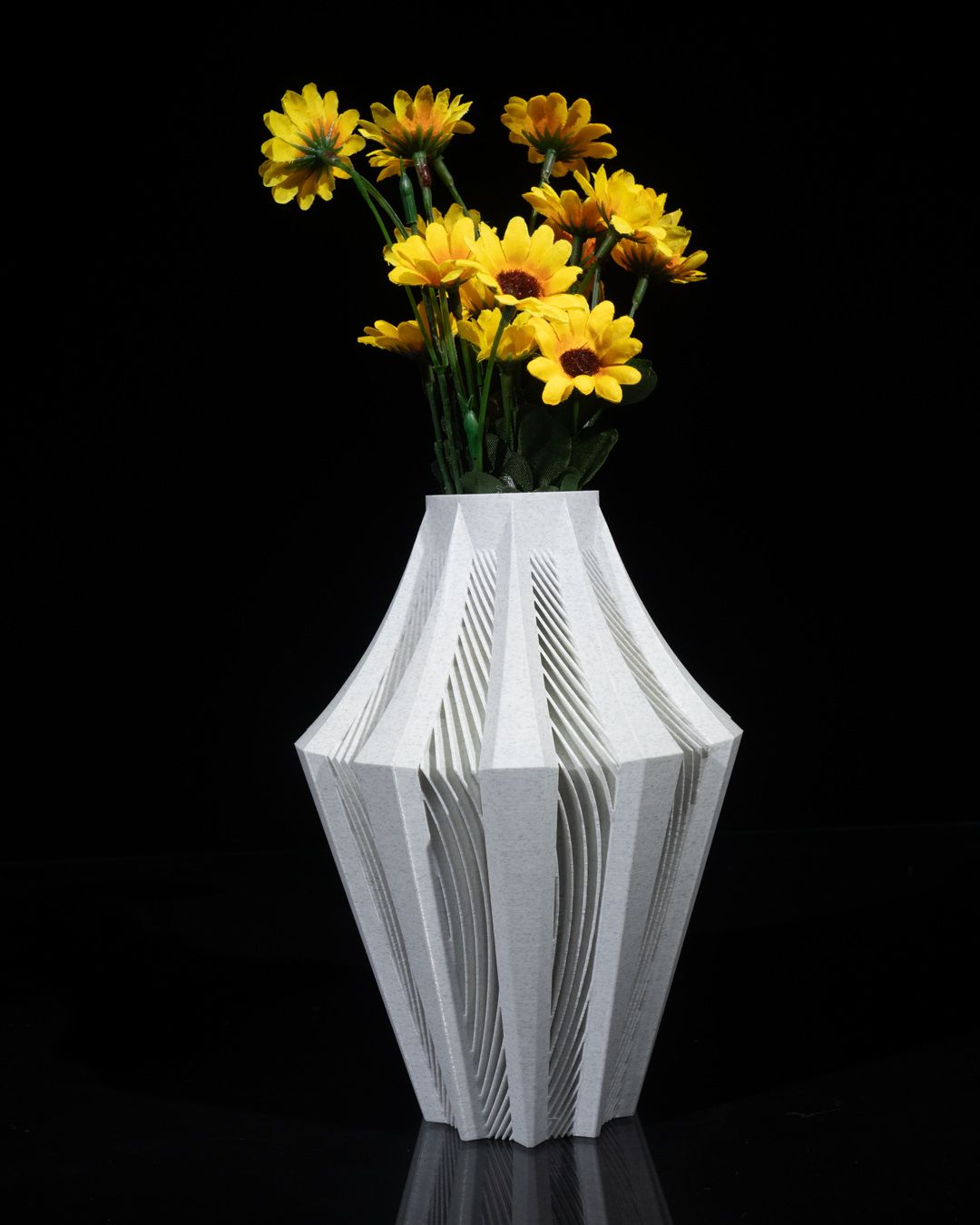 A white, textured vase holds a bouquet of yellow daisies.  The vase is intricately patterned, sits on a dark surface, and the background is black.