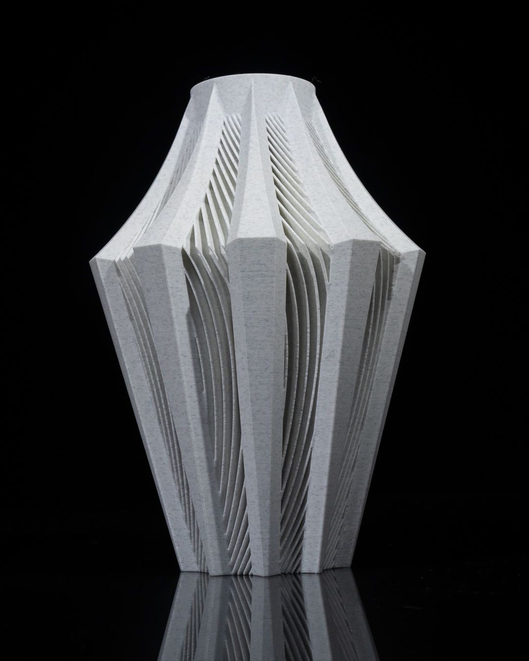 A light grey vase, featuring an intricately carved, pleated design, sits on a reflective black surface.