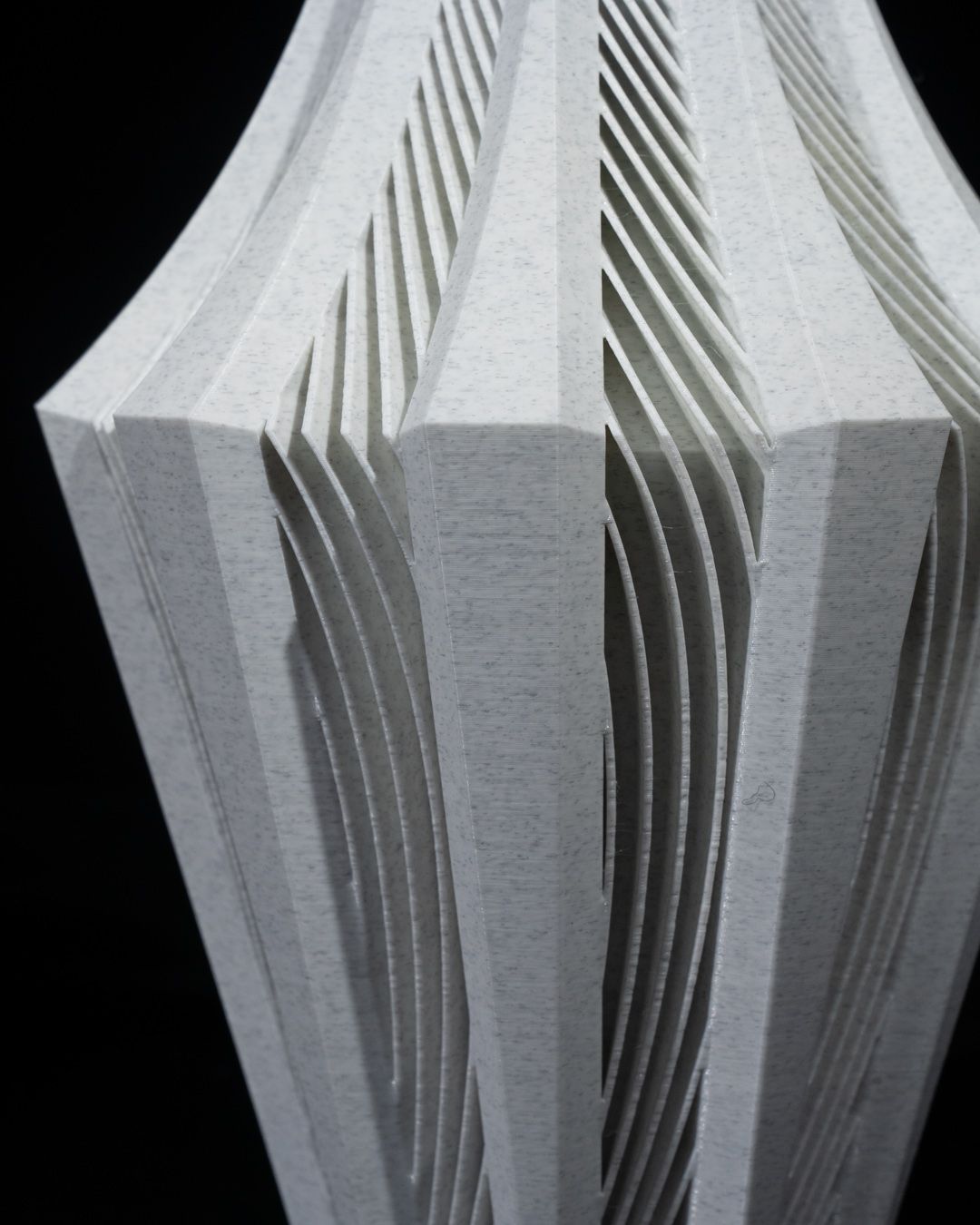 A light grey, intricately carved vase stands, exhibiting a complex pattern of layered, curved lines against a black background.