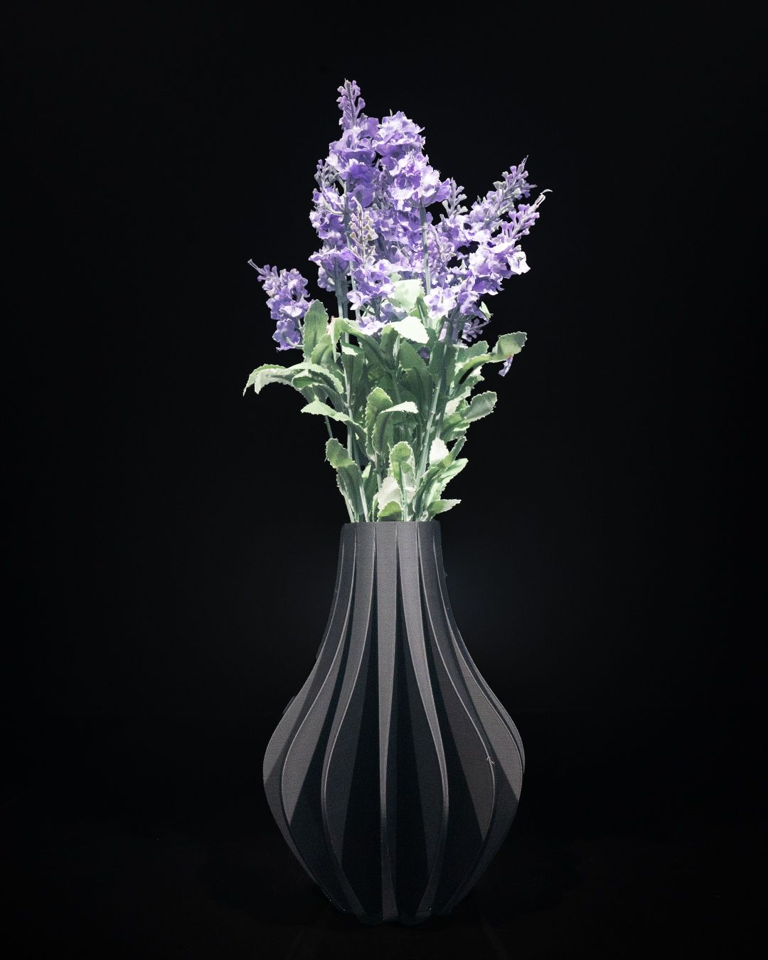 Purple flowers are arranged inside a black vase with vertical ridges, standing on a reflective surface, set against a stark black background.