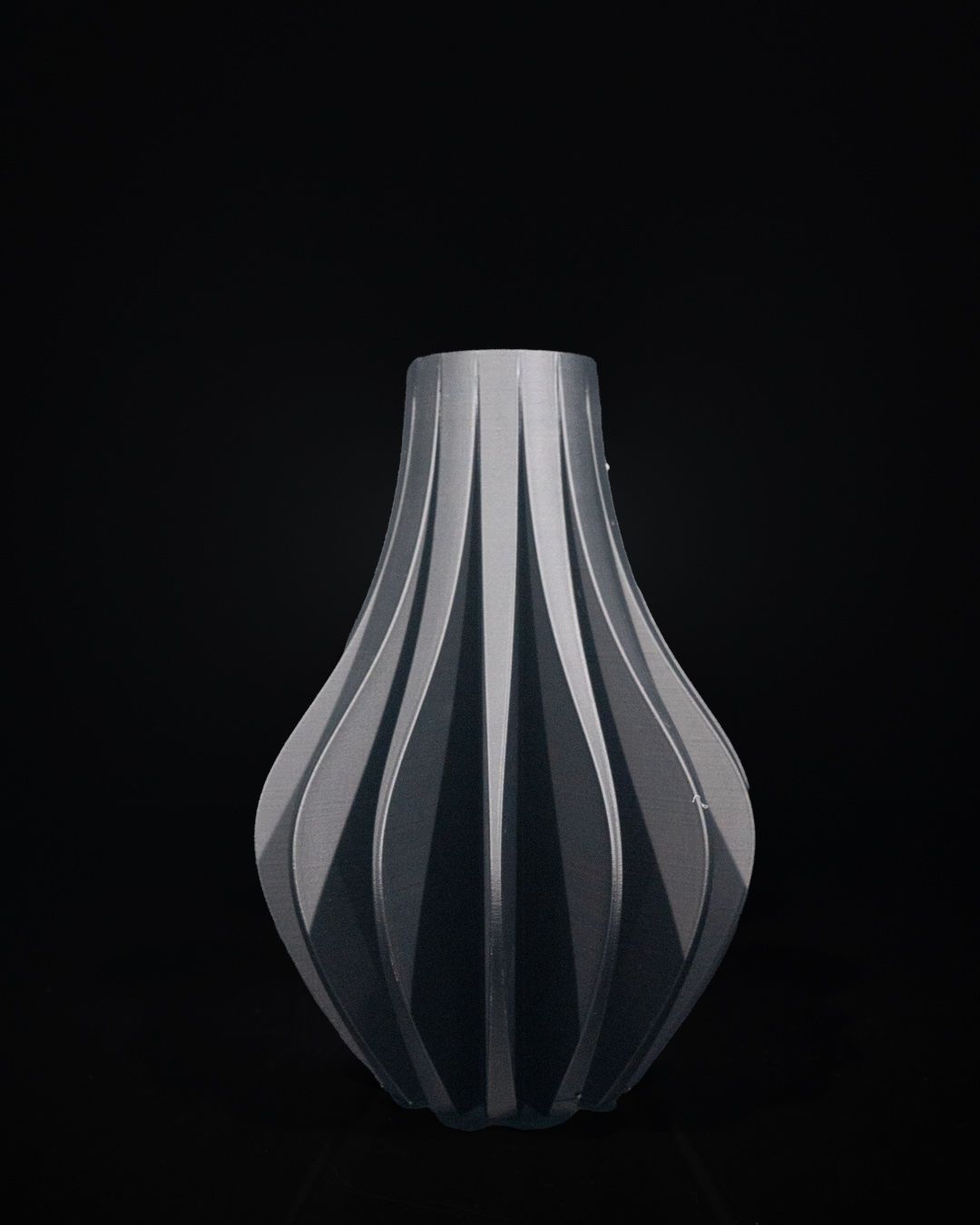 A gray vase with vertical fluting sits stationary against a stark black background. The vase's shape features a bulbous base tapering to a narrow neck.