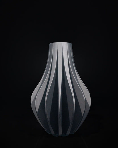 A gray vase with vertical fluting sits stationary against a stark black background. The vase's shape features a bulbous base tapering to a narrow neck.
