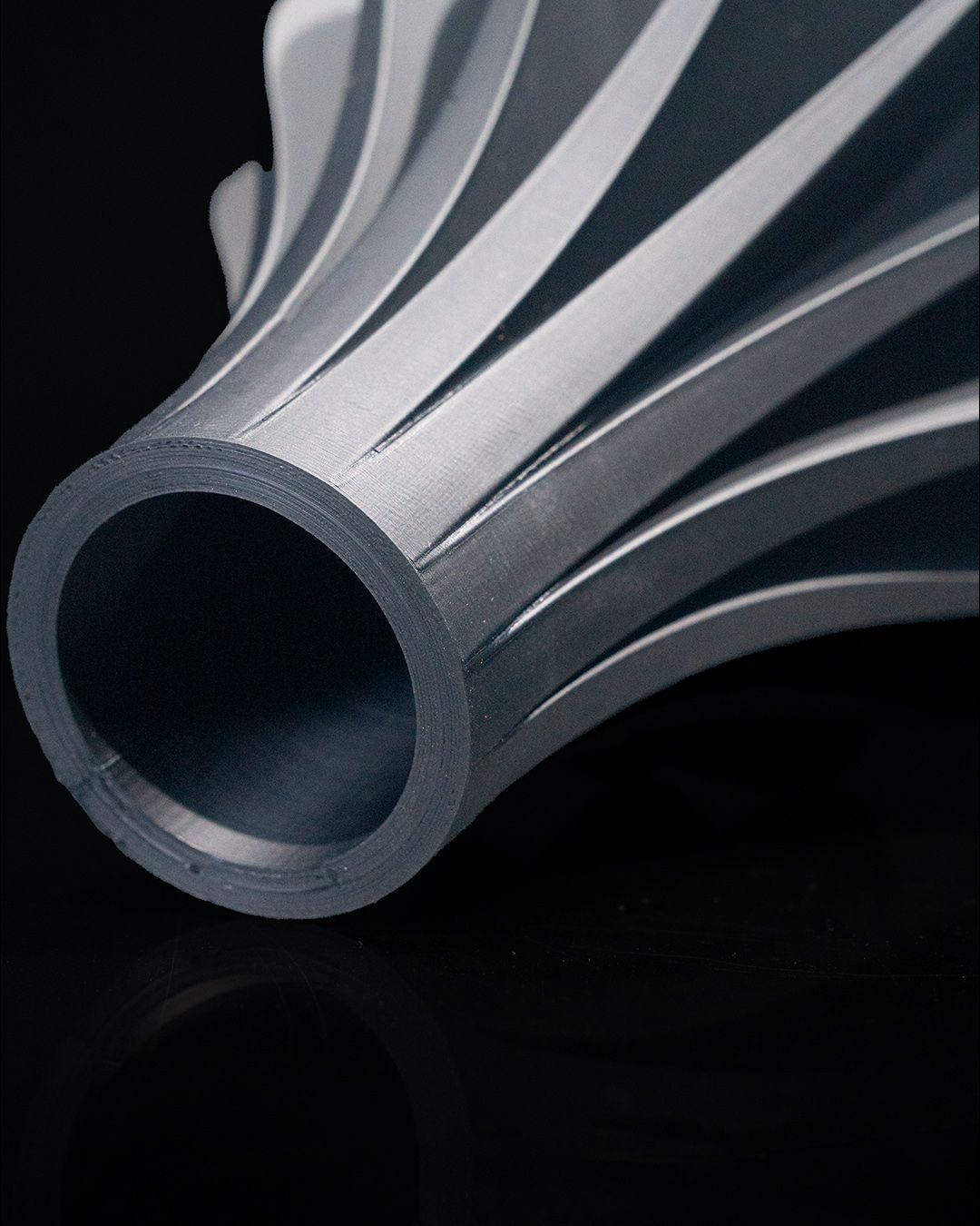 A grey, cylindrical vase flares out with spiraling ridges, resting on a reflective black surface.