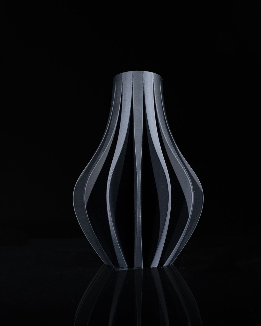 A sculptural vase-like object made of vertical gray strips stands on a reflective black surface against a solid black background.