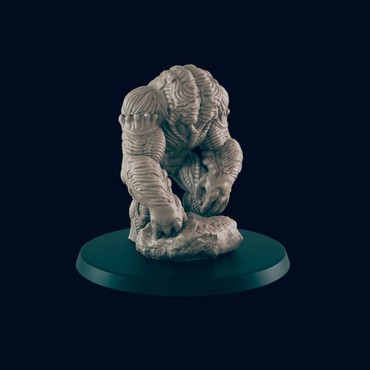 A monster-like, grey miniature figure kneels with hands on a stone, which is resting on a green circular base, against a black backdrop.