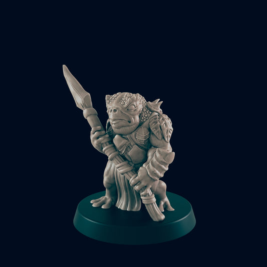 A light-grey amphibian-like humanoid mini-figure stands holding a spear in its left hand. It is on a dark teal base against a dark blue background.