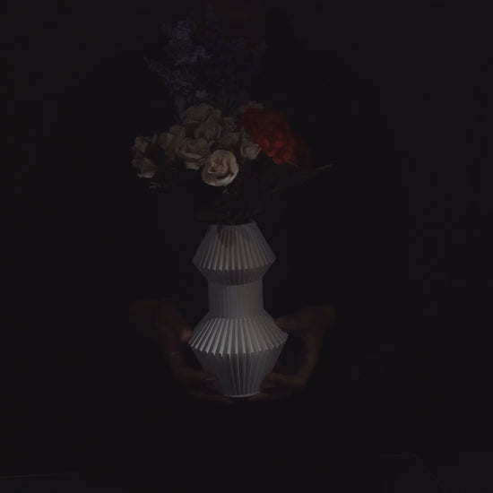 Flowers sit in a white, geometric vase, centered against a black backdrop. The bouquet includes roses, yellow flowers, and tall stalks of wheat.