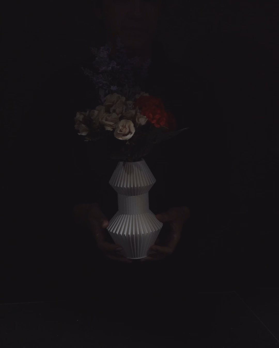 Flowers sit in a white, geometric vase, centered against a black backdrop. The bouquet includes roses, yellow flowers, and tall stalks of wheat.
