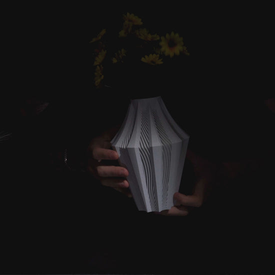 A light-grey, textured lampshade, featuring a geometric, pleated design, stands against a black background.