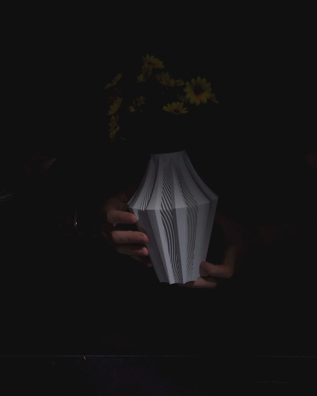 A light-grey, textured lampshade, featuring a geometric, pleated design, stands against a black background.