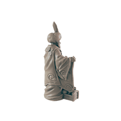 A sculpted figurine of a robed figure sits, appearing to hold a book or scroll, against a black background.