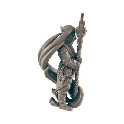 The miniature monk figure, draped with a cape, clutches an ornate staff that extends upwards. The figure is posed against a dark, flat background.
